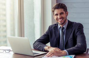 businessman in formal suit using a laptop, looking at camera and smiling while working in office